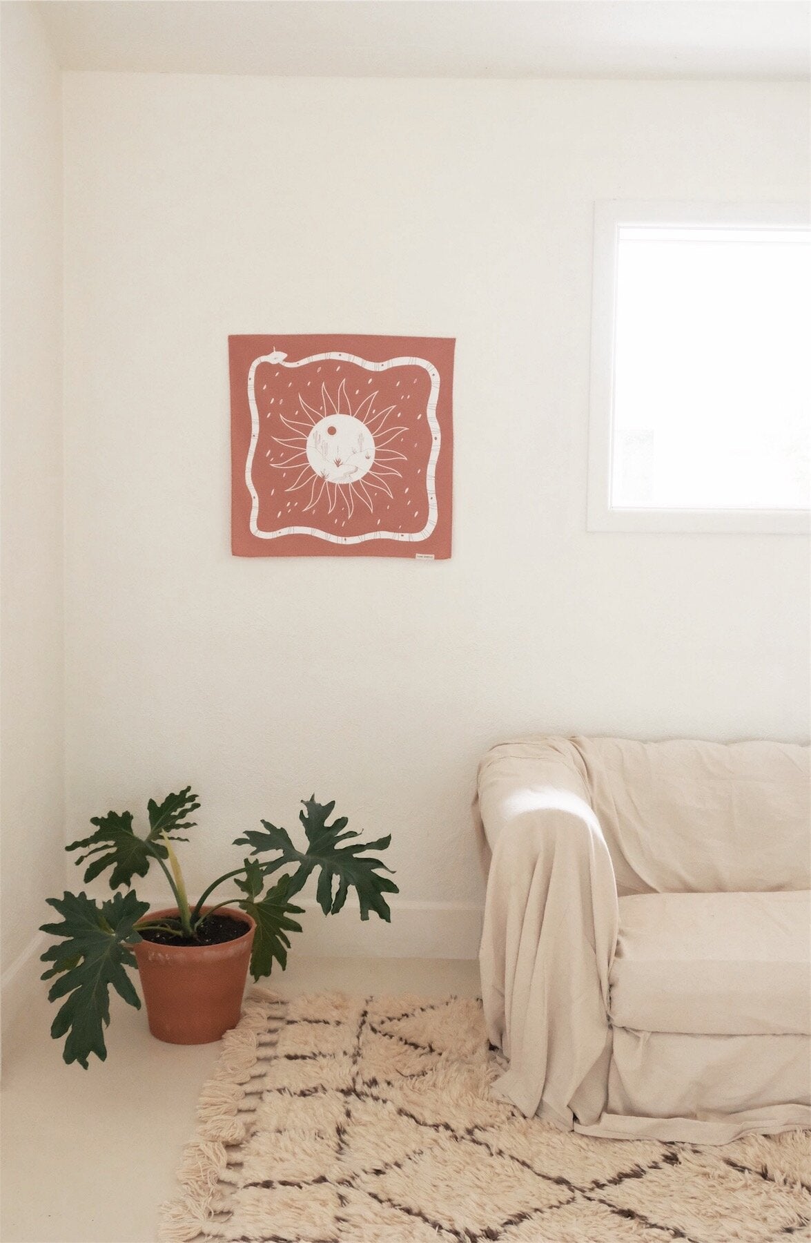 Desert Bandana hanging as a tapestry in a white room with a rug, pretty plant, and white couch. 